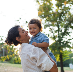 Woman holding a smiling child outdoors in a sunlit park.