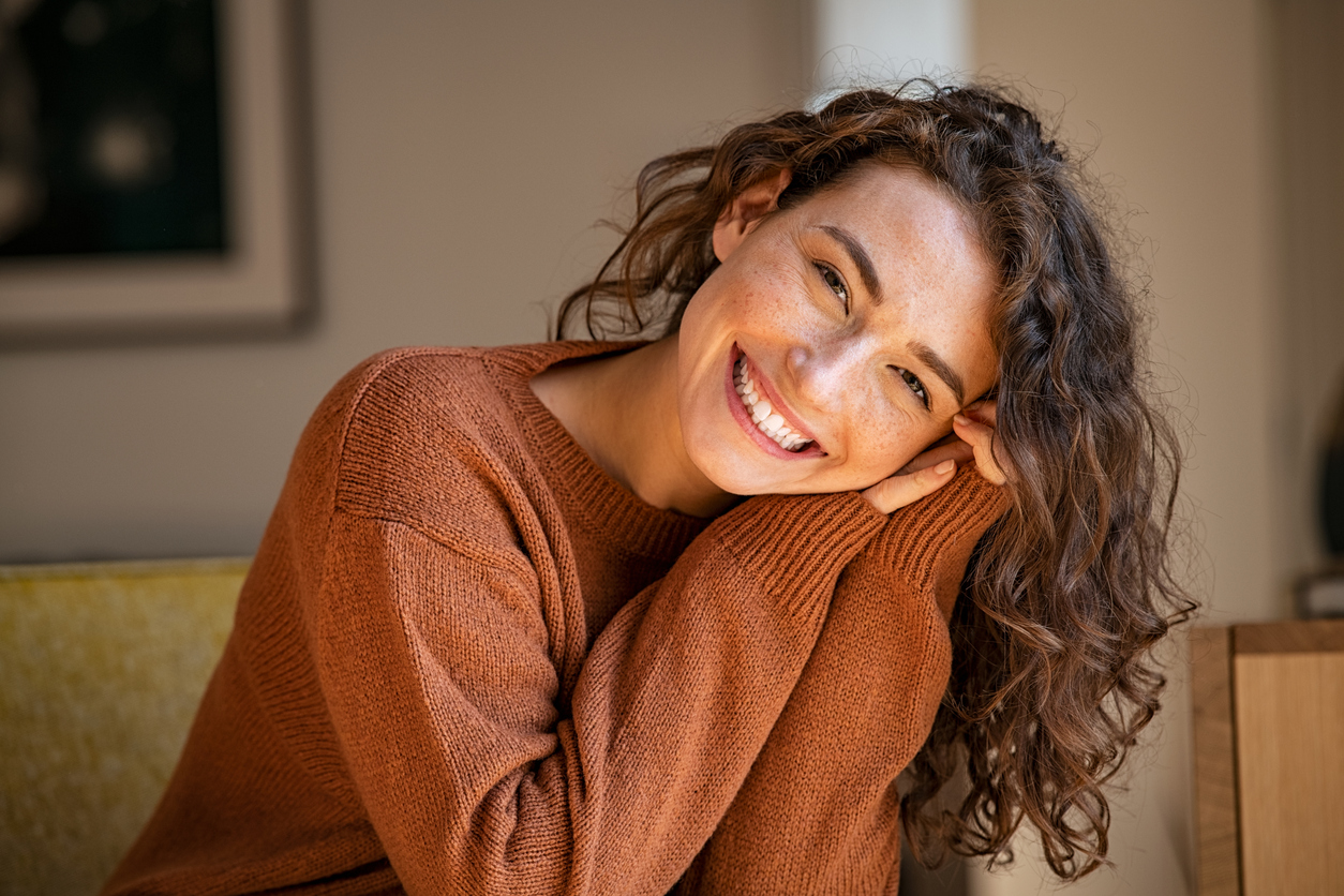 Smiling woman with wavy brown hair in a rust-colored sweater resting her head on her hands indoors.