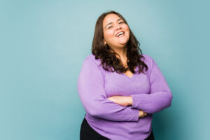 Smiling woman with long dark hair in a purple sweater, arms crossed, against a teal background.