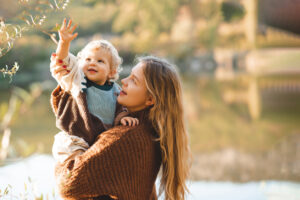 Mother holding a smiling toddler by the water, reaching toward leaves on a branch in a sunlit outdoor setting.