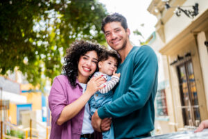 Happy couple holding their baby outdoors on a sunny city street, smiling at the camera.