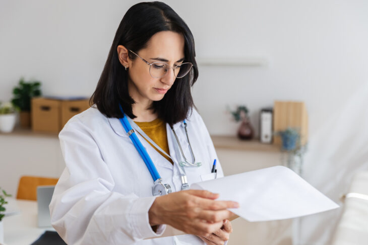 Female doctor in a white coat with a stethoscope reviewing papers at a desk.
