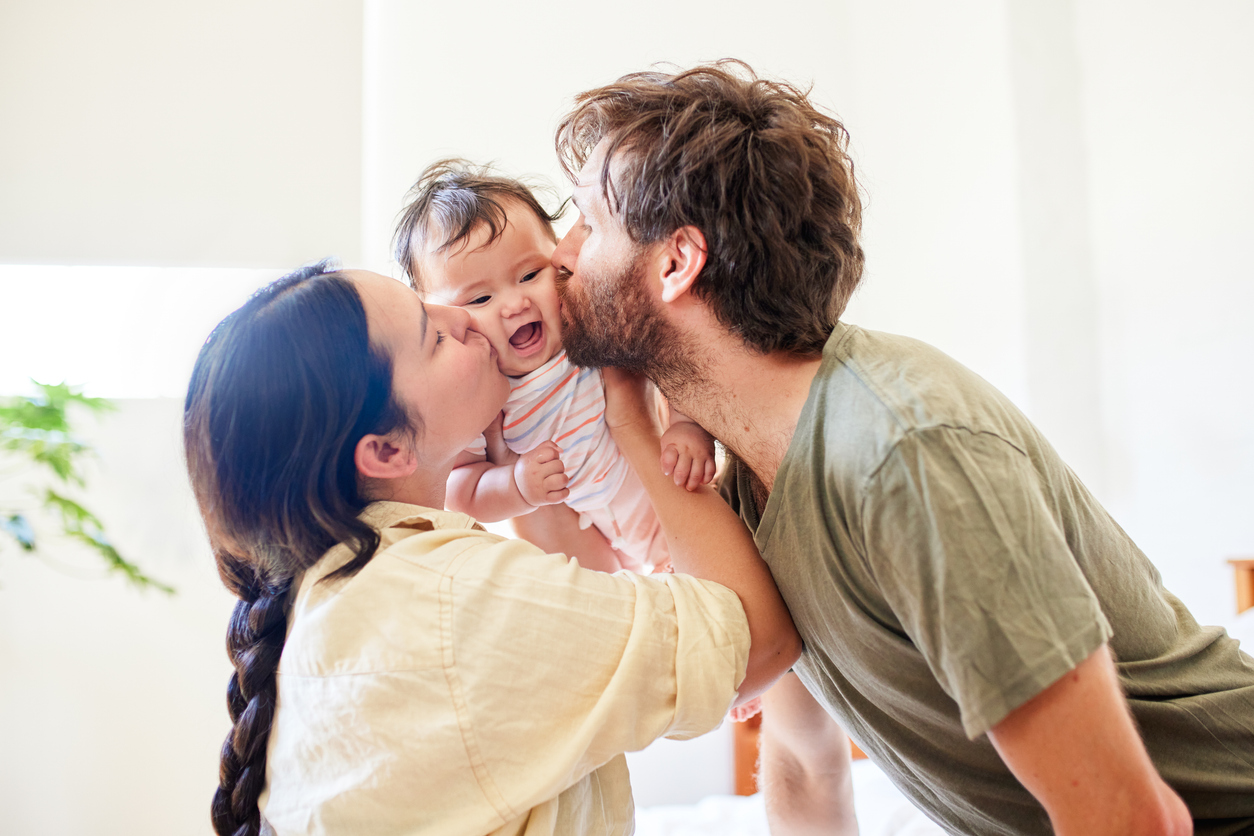 Mother and father kiss their baby, who smiles and is held between them in a bright room on a happy moment.