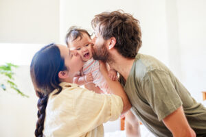 Parents kiss their smiling baby as they lift the child between them in a bright room.