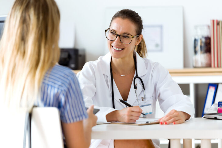 Female doctor in a white coat with stethoscope consulting with a patient across a desk in a clinic, smiling.