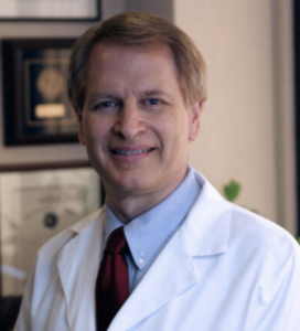 Male doctor in a white coat with a blue shirt and red tie, smiling in a medical office setting.