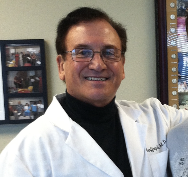 Smiling male doctor in a white coat and black turtleneck, standing in a clinic.