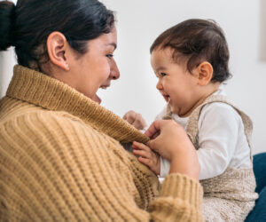 Mother wearing a beige knit sweater smiles at her baby, who playfully grabs her scarf.