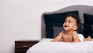 Baby lying on a white bed, propped on elbows, looking up and smiling.