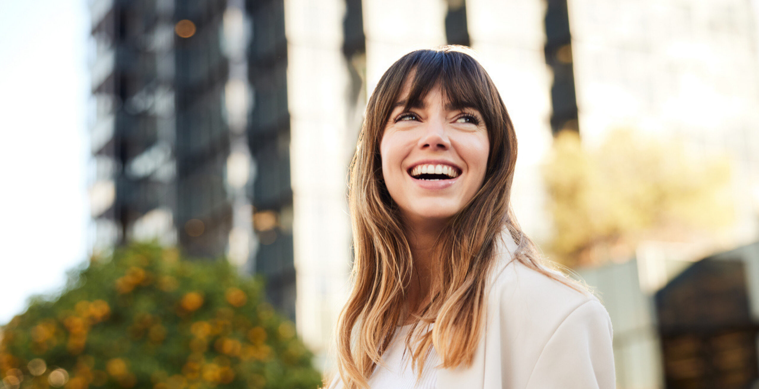 Smiling woman with long brown hair outdoors in an urban plaza with blurred buildings behind her