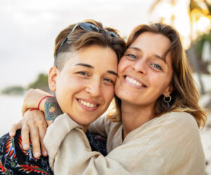 Two women smiling and embracing at a sunny beach.