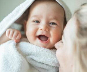 Smiling baby wrapped in a white towel, being kissed by a caregiver.