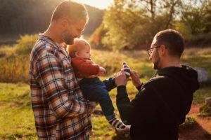 Man in a plaid shirt holds a baby outdoors as another man in a dark jacket shows a striped sock toy in warm sunlight.