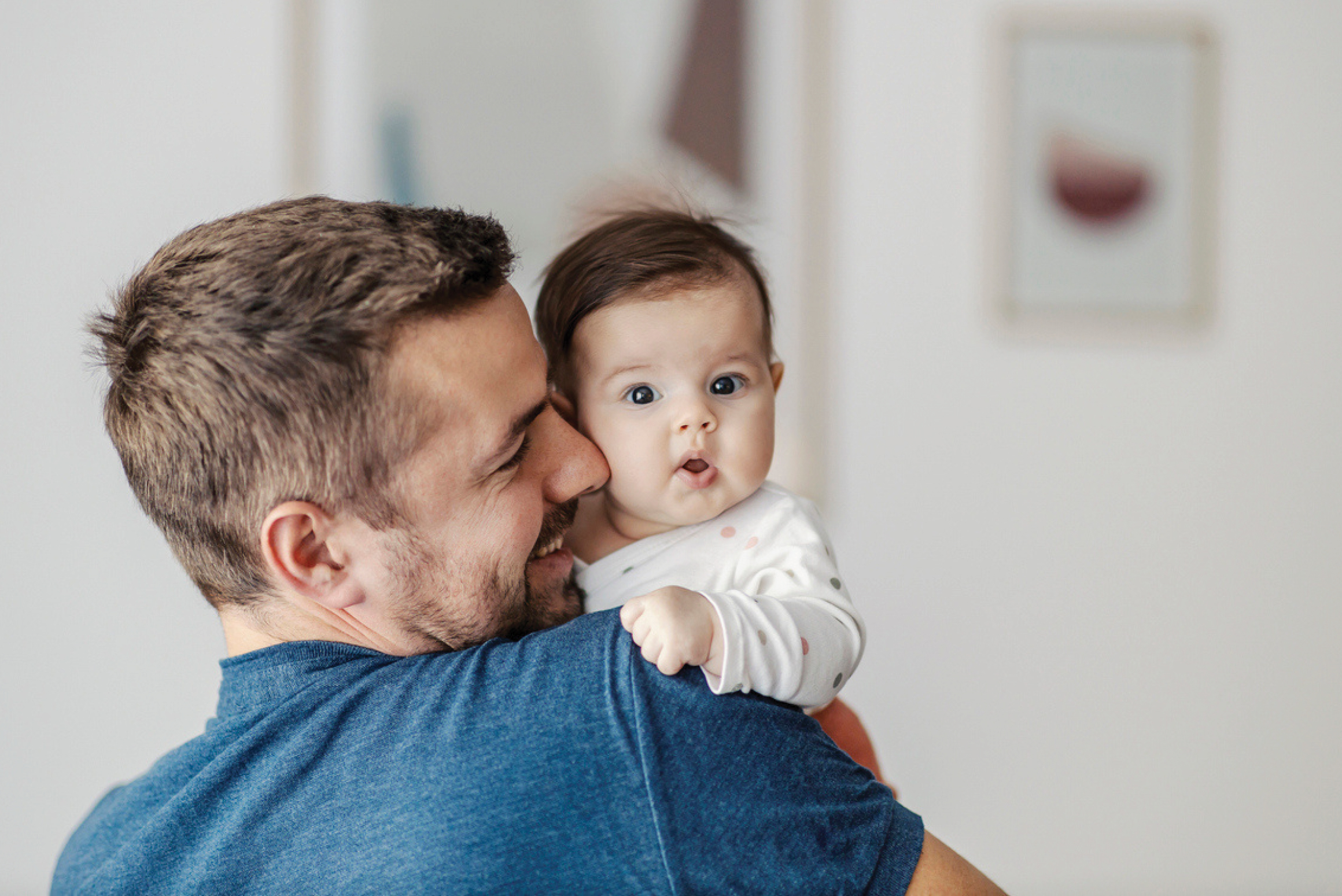 Father in a blue shirt holding a baby close, kissing the baby's cheek in a bright room.