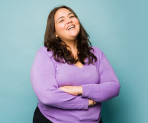Smiling woman with arms crossed wearing a lavender sweater against a teal background.