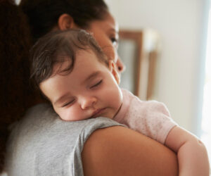 A baby naps peacefully on a parent's shoulder, resting with eyes closed against gray clothing in a cozy indoor setting.