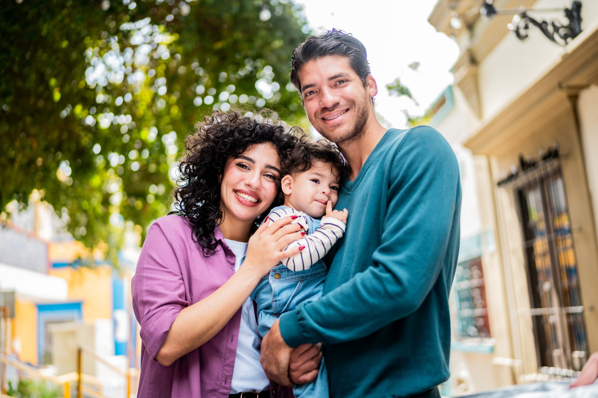Smiling family of three embracing outdoors on a sunny urban street with green trees in the background: mother, father, and baby.
