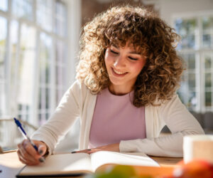 Woman with curly hair smiling as she writes in a notebook at a sunlit desk in a cozy room.