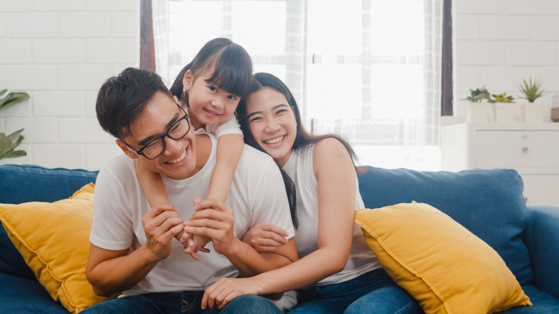 Smiling family of three on a blue couch, daughter hugging dad from behind in a bright living room with yellow pillows.