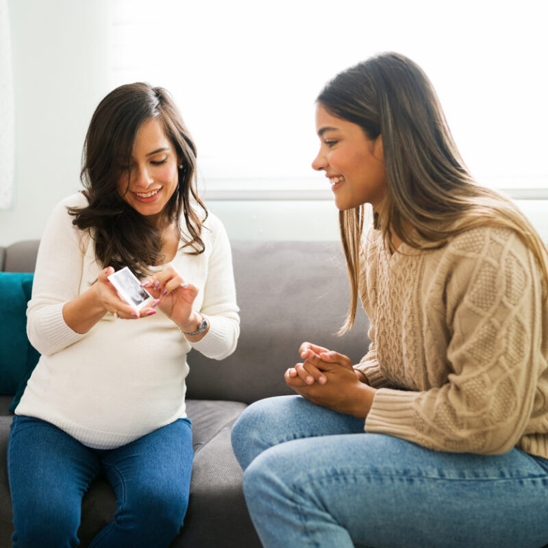 Pregnant woman showing ultrasound photo to friend on a gray sofa at home.