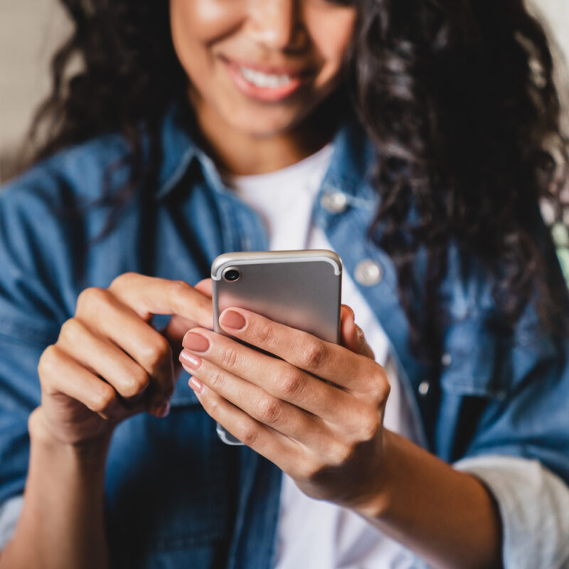 Woman with dark hair in a denim shirt smiling while using a smartphone.