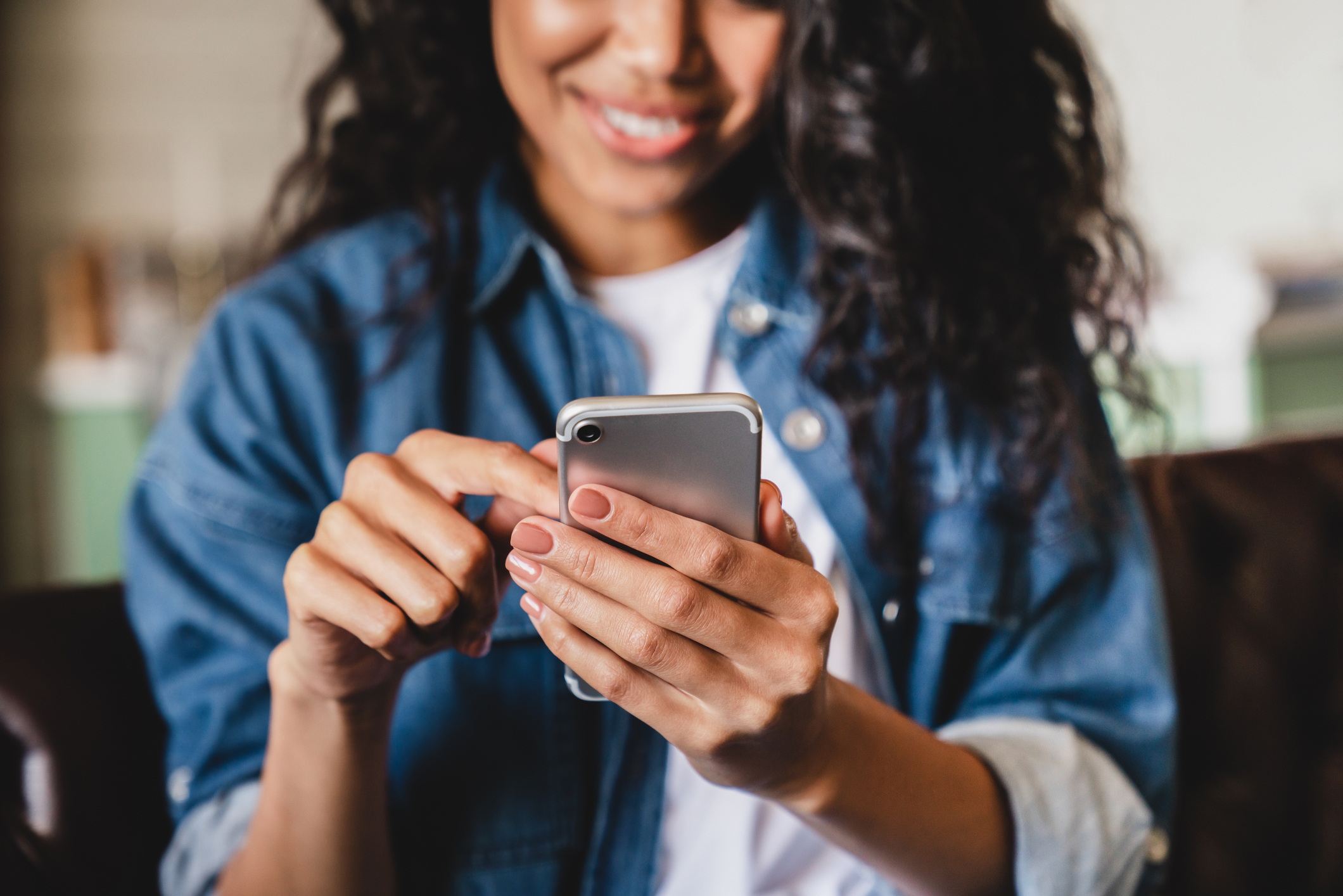 Woman with dark hair in a denim shirt smiling while using a smartphone.