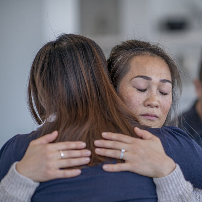 Two people embrace in a supportive hug; the person facing the camera has closed eyes and freckles on her cheeks l.