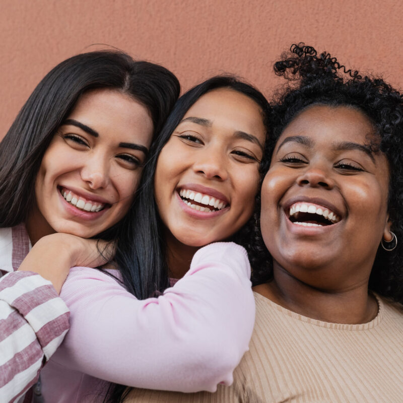 Three smiling women posing for a selfie together against an orange wall.