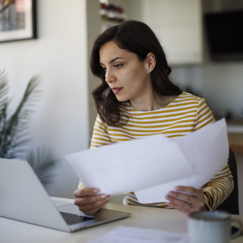 Woman in a yellow-and-white striped shirt sits at a white desk, reviewing papers with a laptop nearby.