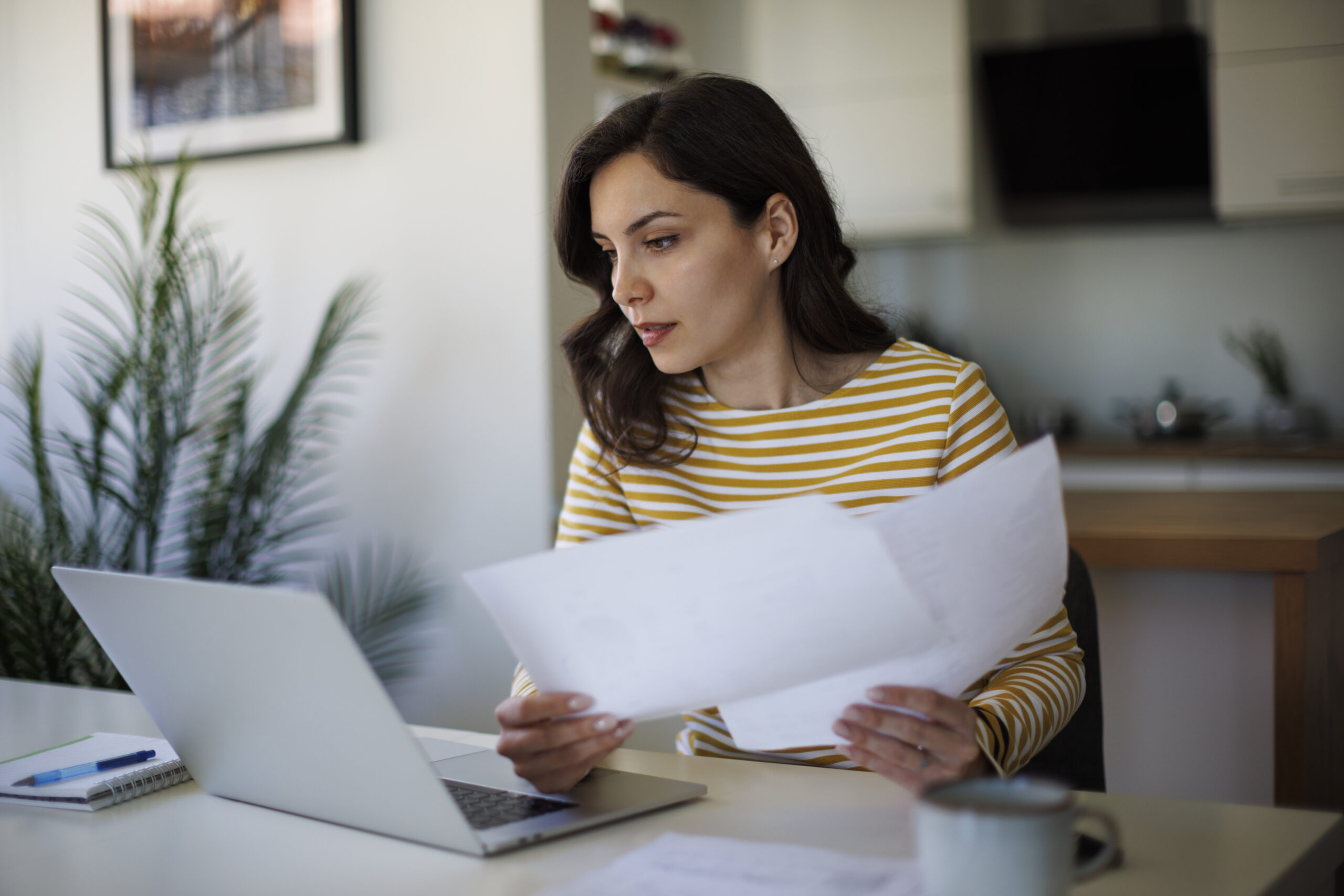 Woman in a yellow-and-white striped shirt sits at a white desk, reviewing papers with a laptop nearby.