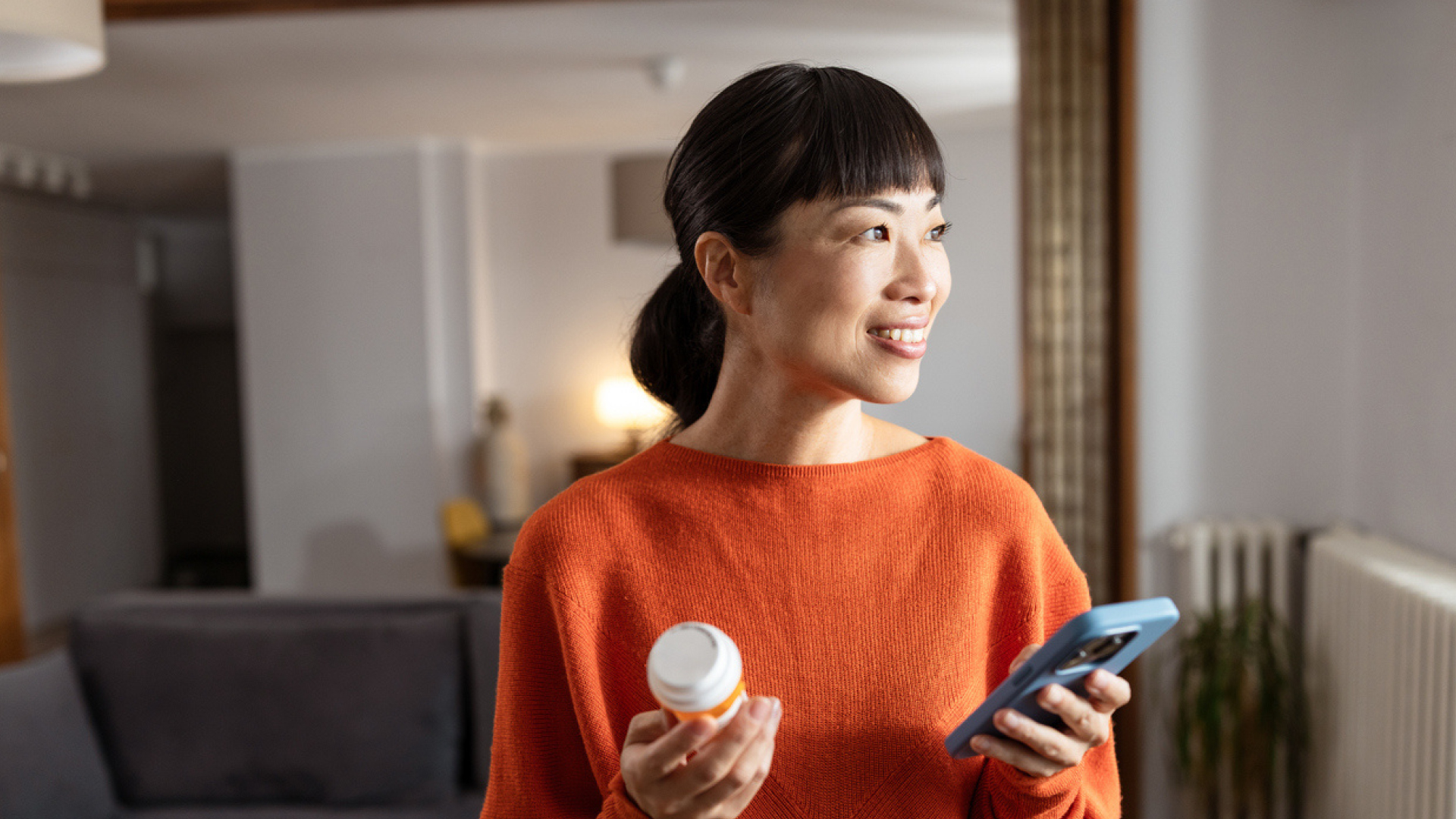 Asian woman in an orange sweater holding a pill bottle and a smartphone, smiling in a living room.